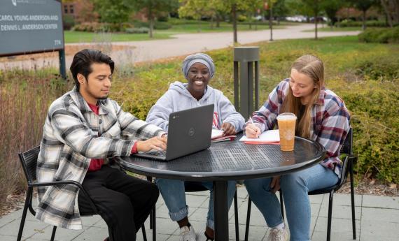 Three undergraduate students sitting outside at table on Hamline University campus, one with laptop 