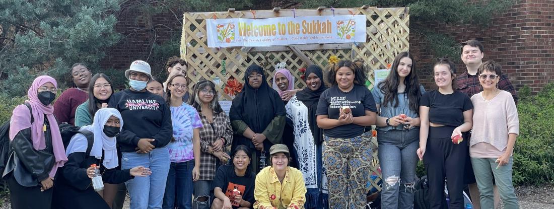 Students stand in front of a structure that says "Welcome to the Sukkah"
