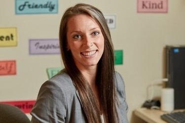 Image of Hamline Alumni student Katie Roselle smiling in the picture using a gray suit in a classroom school in the background.
