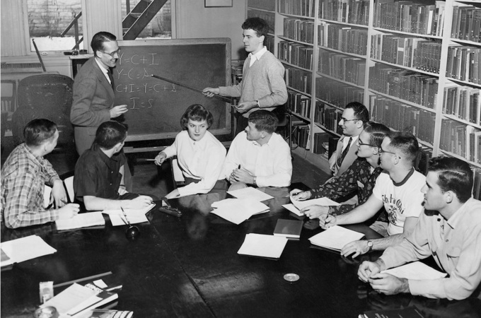 Students and a professor in class, with a student pointing toward a blackboard