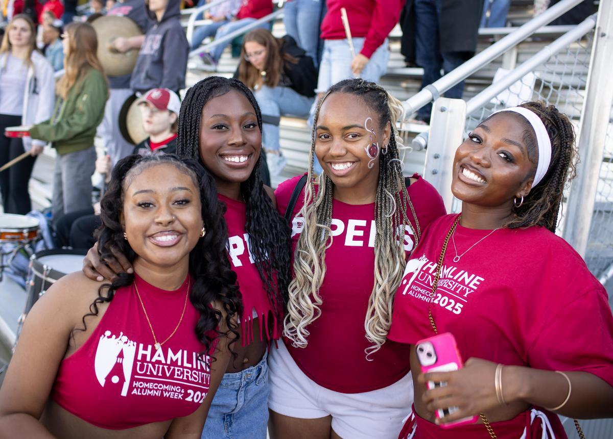 Four young women wearing Hamline t-shirts in front of stands during Homecoming and Alumni week