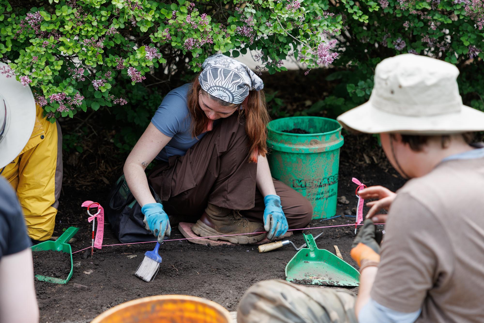 Students collecting artifacts on a dig, sitting beneath a bush