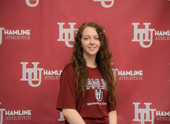 Sydney McCutchen smiling and wearing a Hamline t-shirt in front of a wall covered in Hamline logos