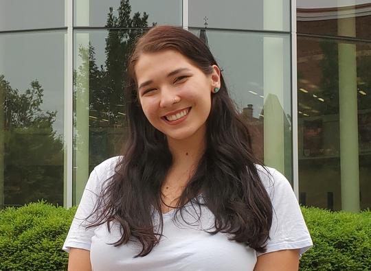 Hamline student Jessie Luévano sitting in front of Anderson Hall smiling at the camera