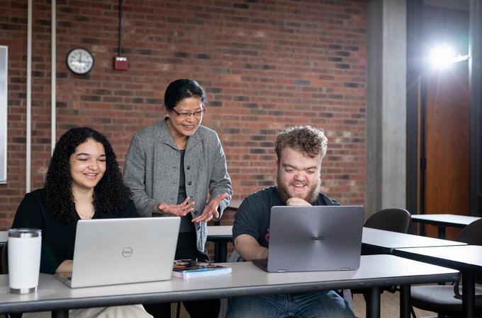Hamline professor stands behind two students who are smiling and working on their laptops