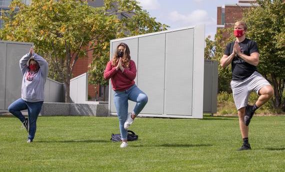 Three Hamline students wearing face masks and holding a yoga tree pose outside on campus.