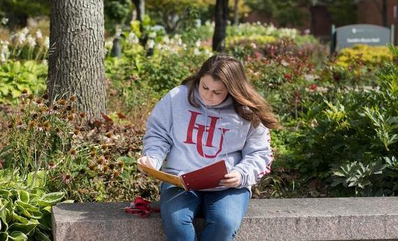 A student in an HU sweatshirt sitting outside reading from their notebook