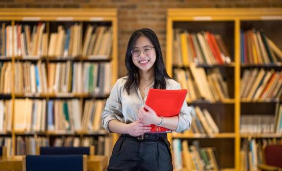 A Hamline student standing in the library holding a folder and smiling