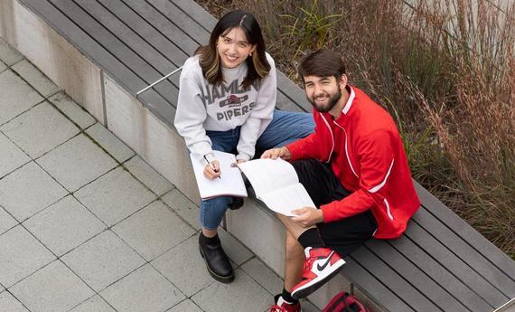 Two Hamline students sitting on a bench outside looking up at the camera smiling