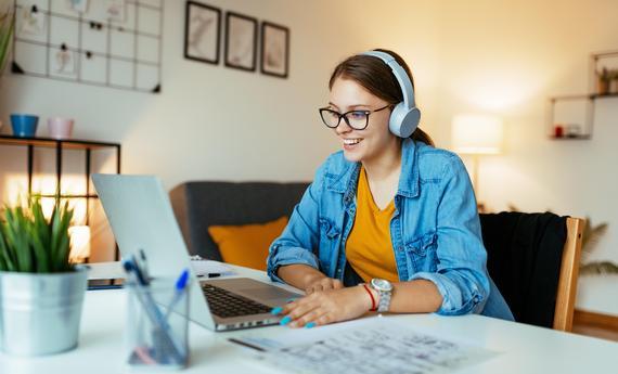 women sitting at desk in front of laptop and smiling at it while wearing headphones