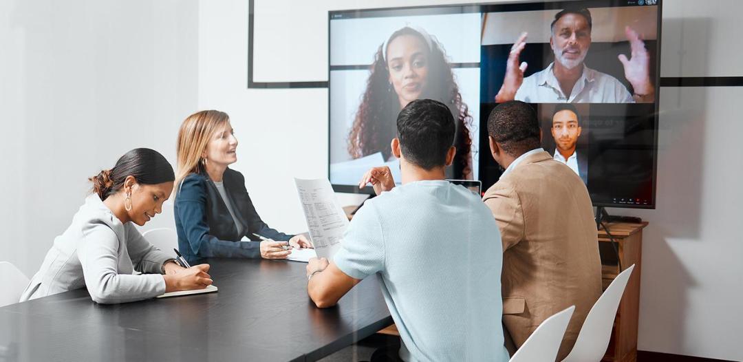 A group of people sitting at a table having a video conference on a large screen