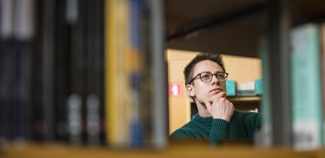 An individual wearing glasses and peering at a library shelf with their hand on their chin