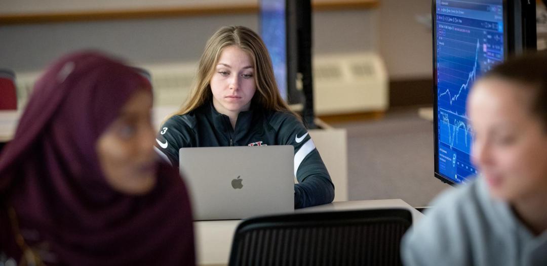 Student working on computer in background, next to a computer display with a graph, data. In front is a woman with a hajab. Business computer lab at Hamline University.