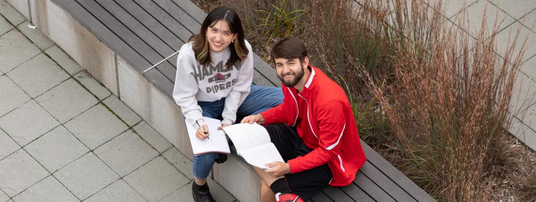 Two Hamline students sitting outside on campus with books and smiling up at the camera