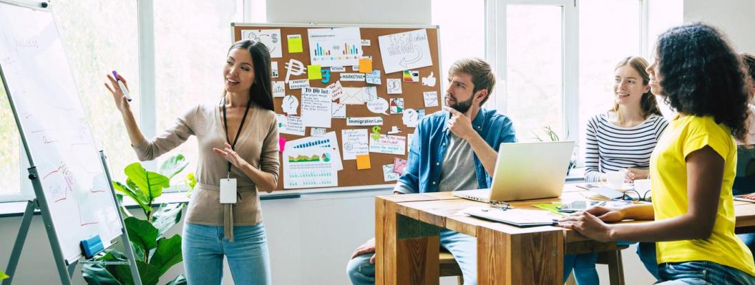 A person gesturing to a whiteboard in front of a table of other people