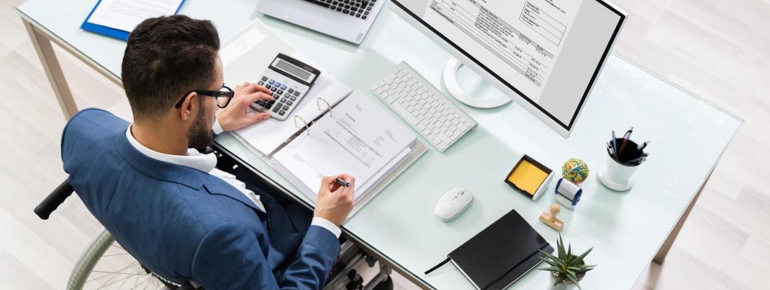 A person sitting at a desk with a computer and calculator writing in a notebook