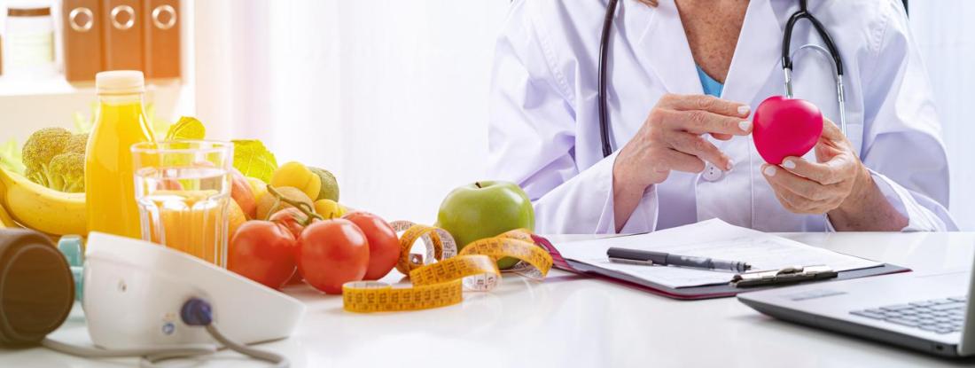 A person in a white coat sitting at a table with many fruits and a laptop