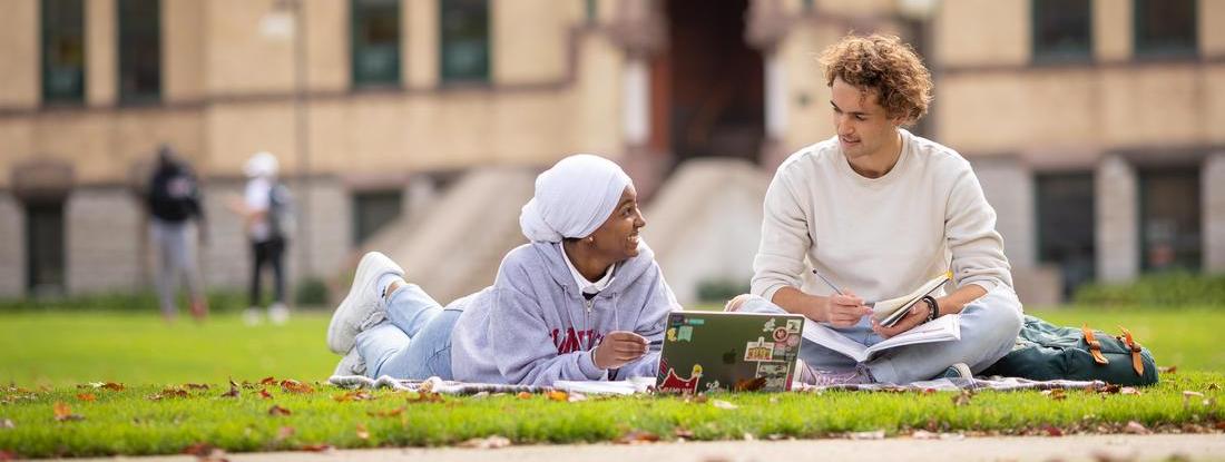 Two Hamline Students looking at a computer in the grass in front of the old main building