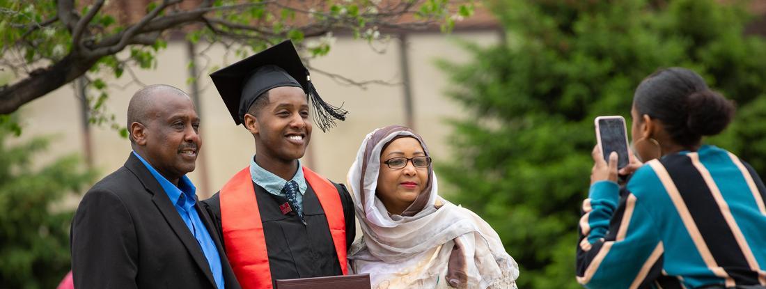 Hamline student in his graduation robes posing with his parents at commencement