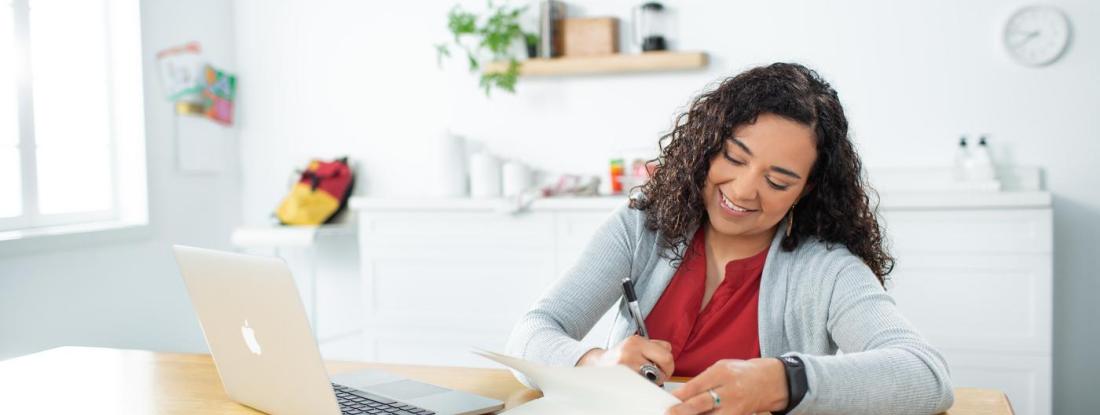 A smiling person sitting at a table working with a laptop and notebook
