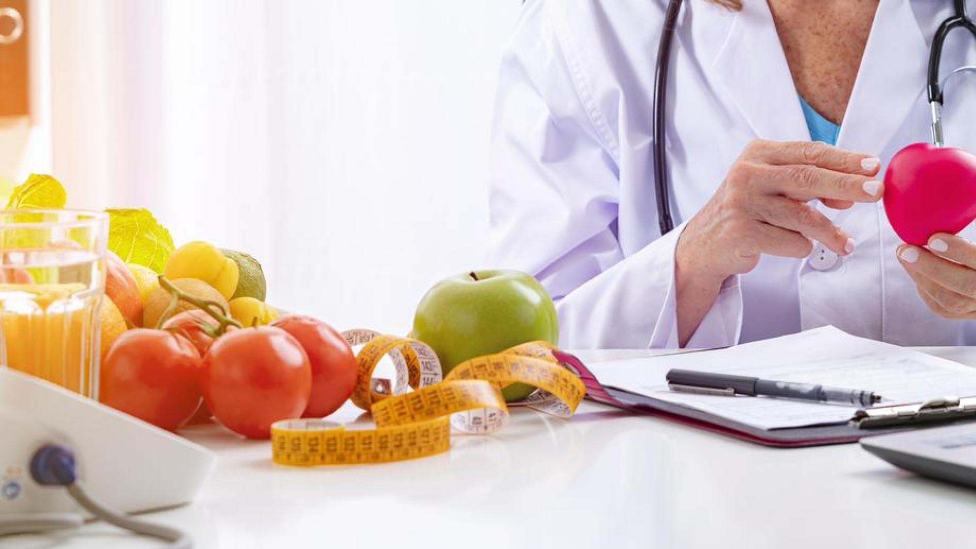 A person in a white coat sitting at a table with many fruits and a laptop