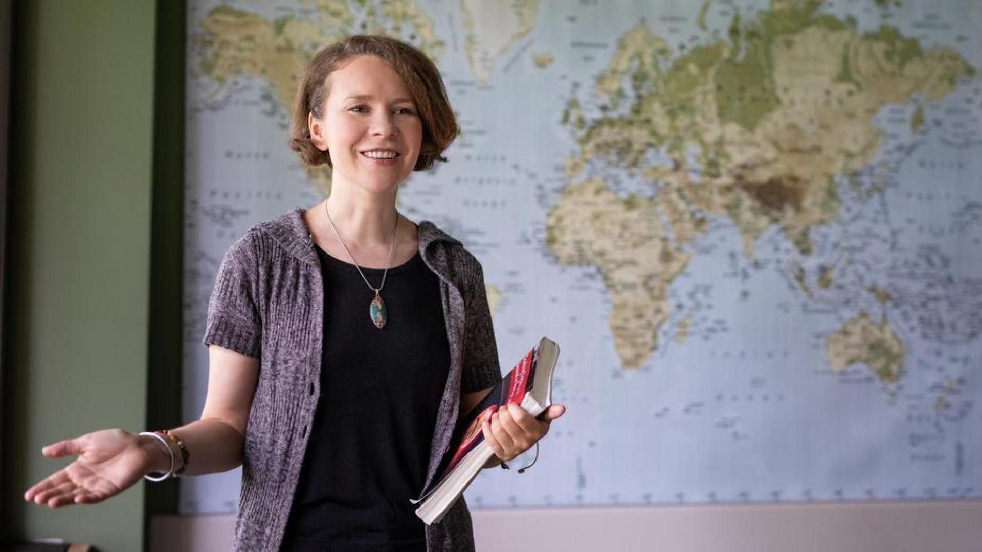 Ilse Griffin holding book and lecturing in a room with a world map behind her 