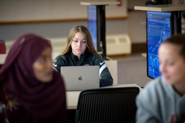 Student working on computer in background, next to a computer display with a graph, data. In front is a woman with a hajab. Business computer lab at Hamline University.