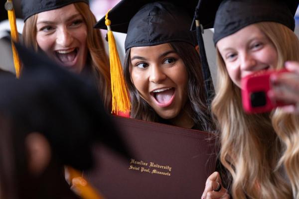 Student with graduation cap holding up diploma and smiling while friend snaps photo